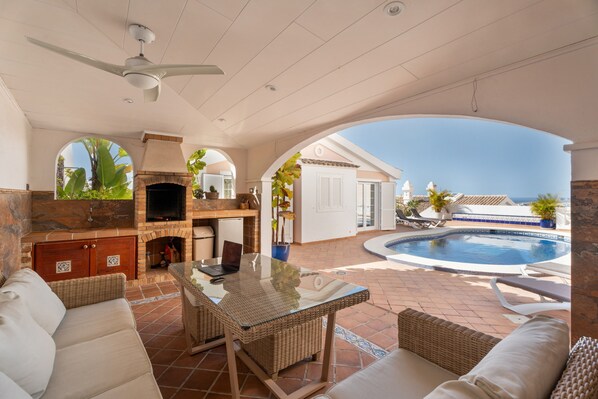 Une terrasse extérieure avec une table en verre, des meubles en osier, une cheminée en briques et une piscine.