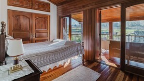 Una camera da letto con soffitto in legno, un letto con copripiumino bianco, un comodino, una lampada e vista su un balcone con ringhiera.