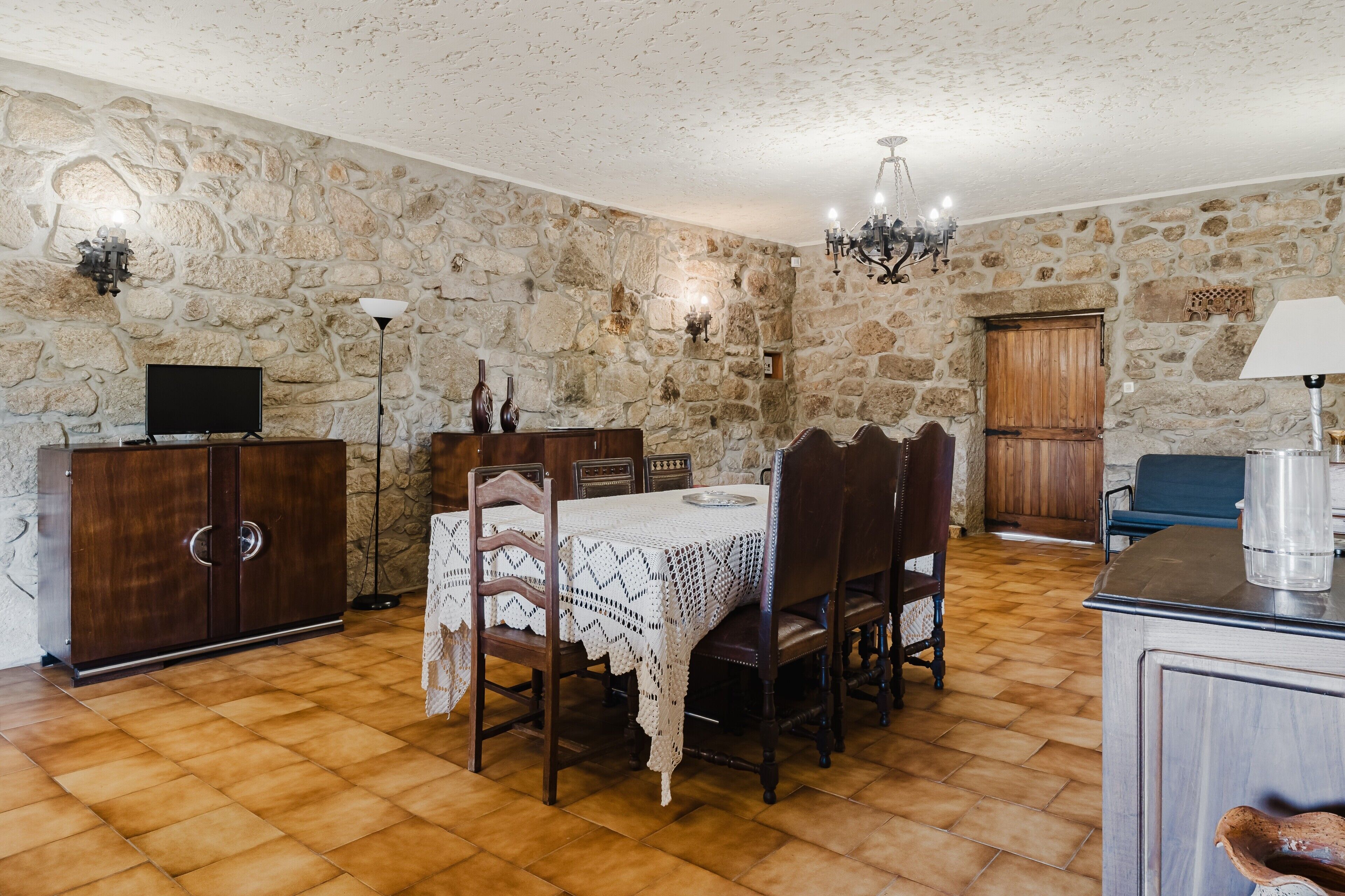 A dining area with a stone wall, a wooden table with a lace tablecloth, chairs, a television on a cabinet, and a chandelier.
