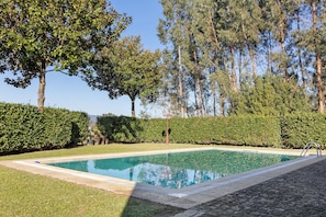 Une piscine rectangulaire entourée d’une terrasse dallée, elle-même bordée d’une pelouse et de haies bien entretenues, avec de grands arbres en arrière-plan.