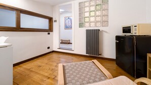 A room with a wooden floor, a small refrigerator, a microwave, a radiator, and a window with frosted glass panels.