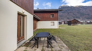Ein modernes Gebäude mit einer Terrassenfläche, die einen Tisch und Stühle bietet, einer Holzfassade und Blick auf eine gebirgige Landschaft.