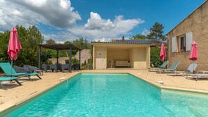 Une piscine avec des chaises longues et des parasols, un espace de détente couvert et un bâtiment doté d’un toit en tuiles.