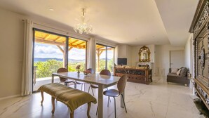 Une pièce spacieuse avec une grande table à manger, des chaises, un banc, un lustre, une cheminée et une vue dégagée sur l’extérieur grâce à de grandes fenêtres.