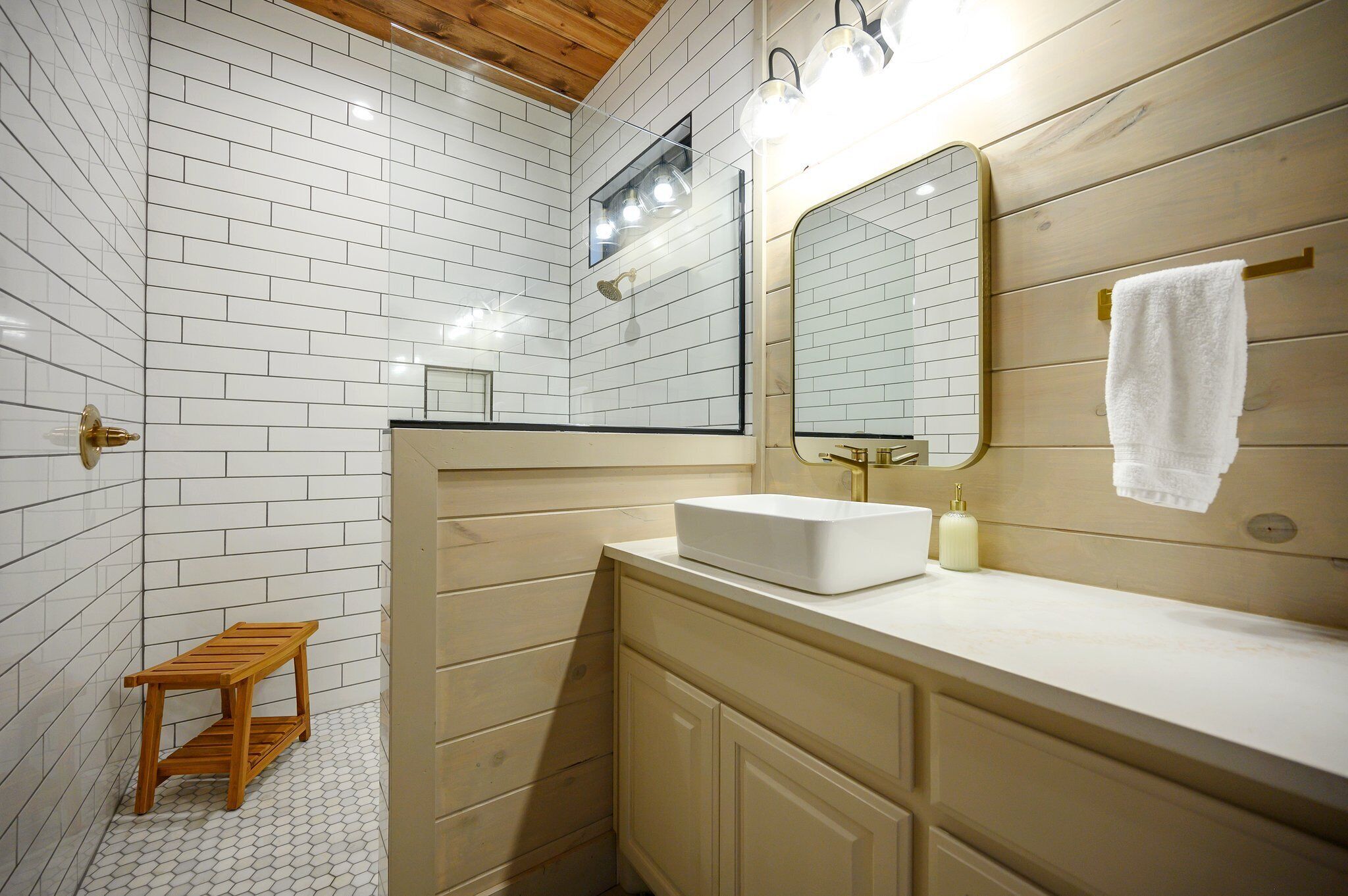 A bathroom with white tiled walls, a wooden vanity, a white sink, a mirror, and a small wooden stool.