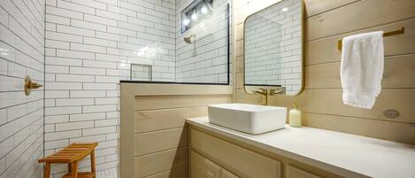 A bathroom with white tiled walls, a wooden vanity, a white sink, a mirror, and a small wooden stool.