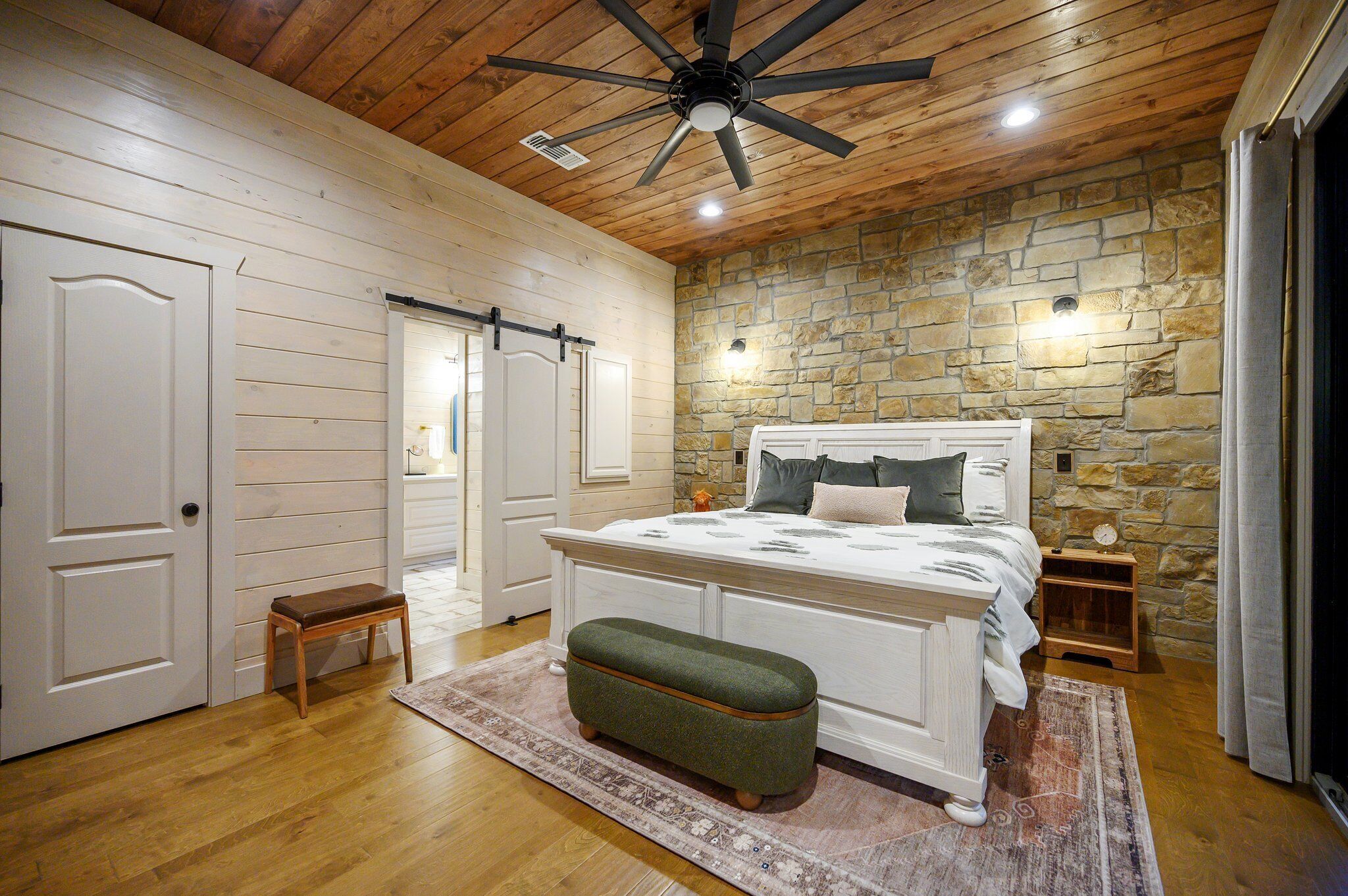 A bedroom with a wooden ceiling, stone wall, and a bed with a white headboard.