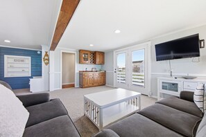A modern living room with a white coffee table, grey sofas, and a kitchen area with wooden cabinets and a built-in oven. - Resting Beach Face: Semi-Oceanfront, Classic Nags Head Flat Top Home, Private Pool, Hot Tub (Nags Head)