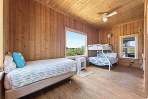 A wooden cabin room with bunk beds, a single bed, a ceiling fan, and large windows. - Resting Beach Face: Semi-Oceanfront, Classic Nags Head Flat Top Home, Private Pool, Hot Tub (Nags Head)