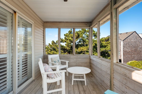 A covered porch with two white chairs and a small table, large windows, and a view of trees and a house. - Hawk's Nest: Semi-Oceanfront, Hot Tub, Close to Beach Access (Nags Head)