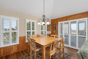 A dining area with a wooden table and chairs, white shutters on the windows, and a chandelier. - Hawk's Nest: Semi-Oceanfront, Hot Tub, Close to Beach Access (Nags Head)