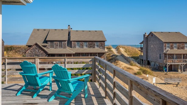 Une terrasse en bois avec deux fauteuils Adirondack turquoise, donnant sur une plage bordée de maisons situées sur les dunes et sous un ciel bleu azur.