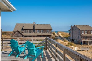 A wooden deck with two turquoise Adirondack chairs, overlooking a beach with dune houses and a clear blue sky. - Hawk's Nest: Semi-Oceanfront, Hot Tub, Close to Beach Access (Nags Head)