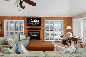 A living room with a fireplace, a television, and a ceiling fan. - Hawk's Nest: Semi-Oceanfront, Hot Tub, Close to Beach Access (Nags Head)