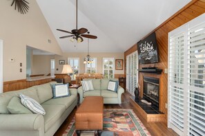A living room with a sofa, ottoman, fireplace, ceiling fan, and a TV mounted on the wall. - Hawk's Nest: Semi-Oceanfront, Hot Tub, Close to Beach Access (Nags Head)