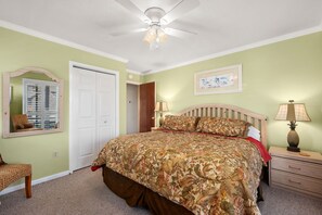 A bedroom with a large bed, a ceiling fan, a nightstand with a lamp, a chair, and a door with a mirror.