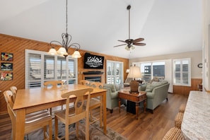 A spacious living room with a wooden dining table, a sofa, and a ceiling fan. - Hawk's Nest: Semi-Oceanfront, Hot Tub, Close to Beach Access (Nags Head)