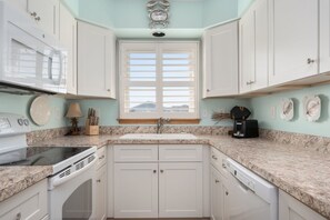 A kitchen with white cabinets, a granite countertop, a built-in oven, a dishwasher, and a window with blinds. - Hawk's Nest: Semi-Oceanfront, Hot Tub, Close to Beach Access (Nags Head)