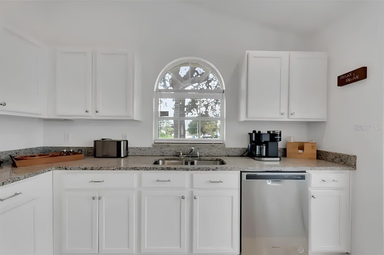 A modern kitchen with white cabinets, a granite countertop, a built-in dishwasher, and a window with a view of trees.
