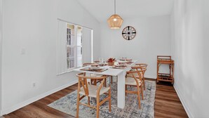 En matplats med ett vitt bord dukat för sex personer, trästolar, en hängande lampa och en väggklocka.