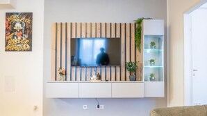 Una sala de estar con un televisor montado en la pared, listones de madera, una estantería con plantas y un póster.