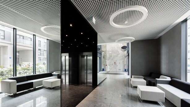 A modern lobby with a central elevator, seating areas, and large windows offering views of the city.