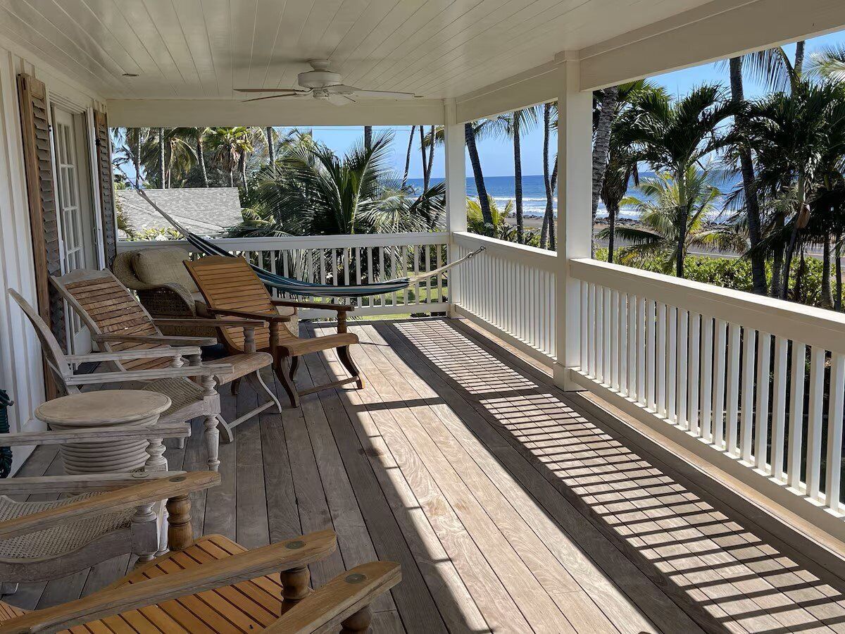 Una terraza de madera con mobiliario exterior, barandal blanco y vista a palmeras y al océano.