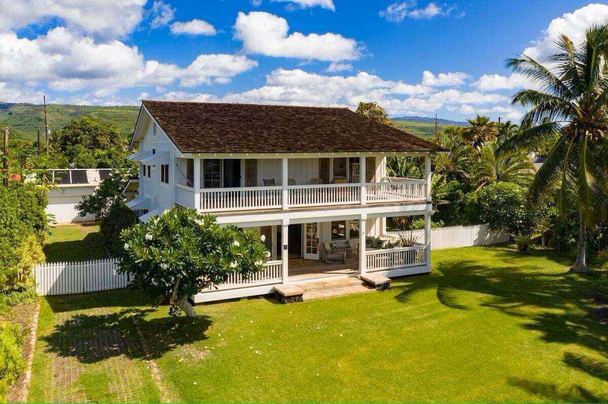 A two-story house with a balcony, surrounded by a well-maintained lawn and palm trees.