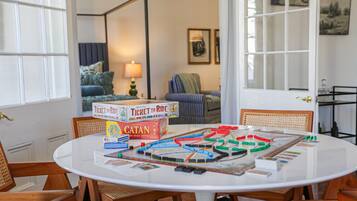 A round table with board games, including Ticket to Ride and Catan, in a room with wooden chairs and a view of a living area with a lamp and framed pictures.