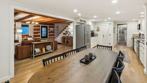 Amplia sala de estar con mesa de comedor de madera, sofá y cocina con armarios blancos y refrigerador.