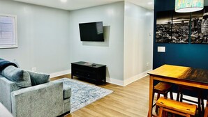 A modern living room with a grey sofa, a wooden dining table with benches, a flat-screen TV mounted on the wall, and a cityscape artwork on the wall.