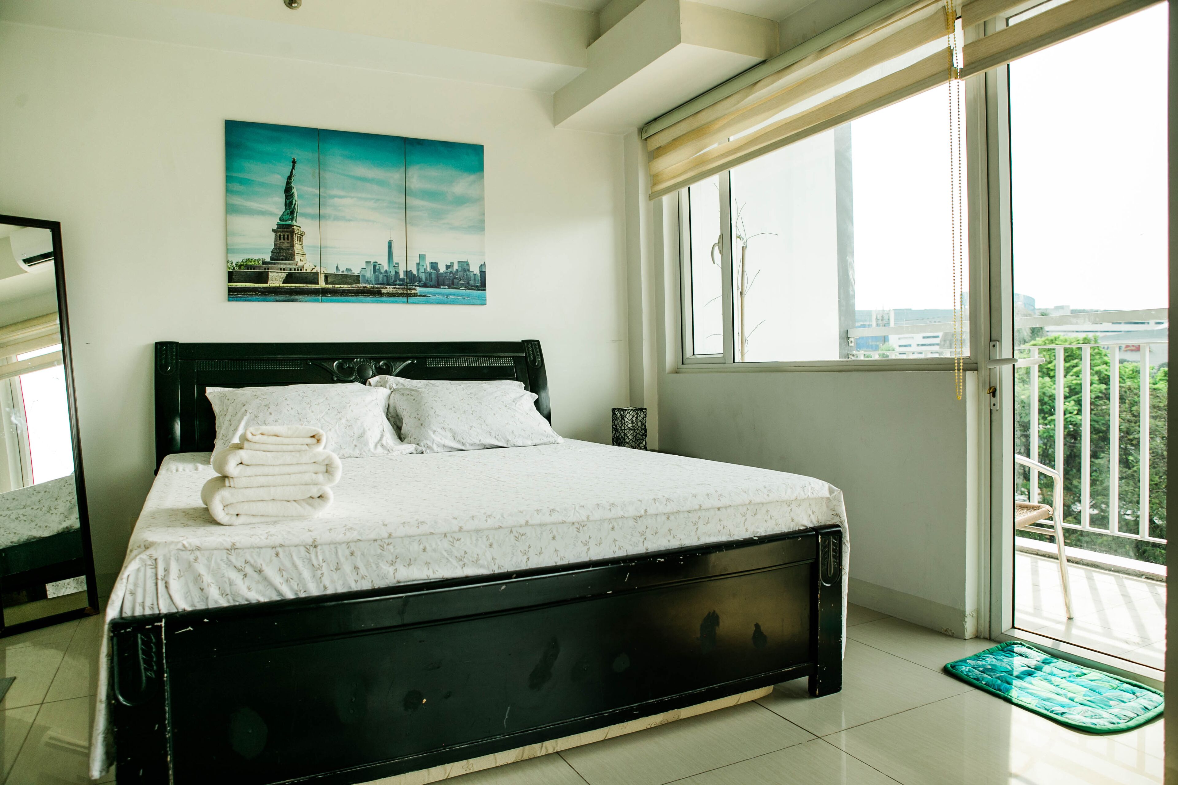 A bedroom with a bed, a window with a view of the city, and a painting of the Statue of Liberty.