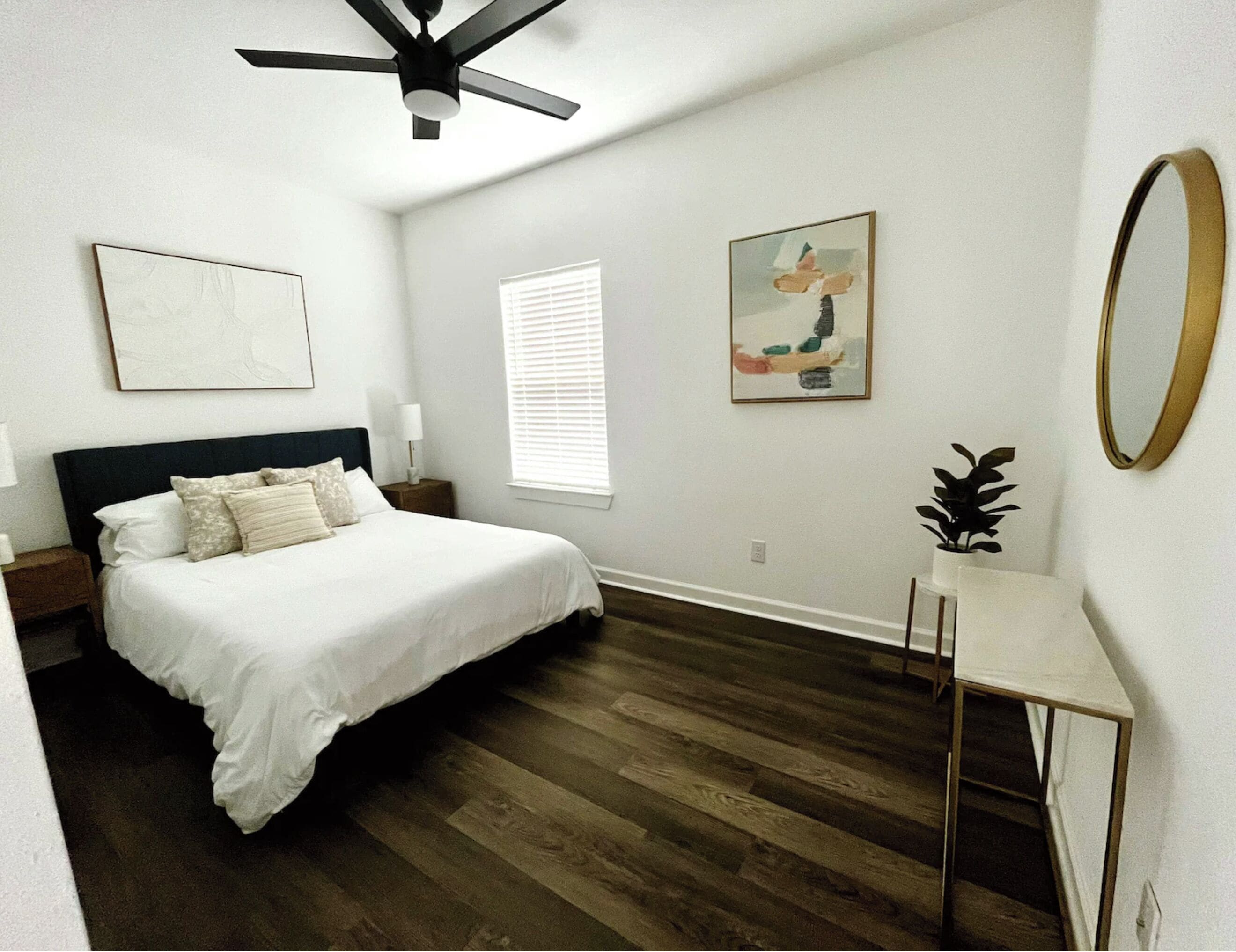 Une chambre à coucher comprenant un lit, un ventilateur de plafond, une table de chevet avec une lampe, une fenêtre avec des stores, un tableau encadré, une petite table avec une plante et un miroir sur le mur.