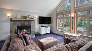 A cozy living room with a fireplace, comfortable seating, and large windows offering a view of the outdoors.