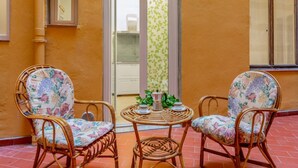 En liten sittplats utomhus med flätade stolar och ett bord, en dörr som leder till ett badrum och ett fönster med utsikt.