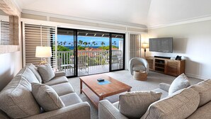A spacious living room with a large sliding glass door leading to a balcony with a view of palm trees and the ocean. There is a flat-screen TV mounted on the wall, a comfortable sofa set, and a wooden coffee table. - Kiahuna Plantation Resort 116 Partial Ocean 2BD 2B (Koloa)