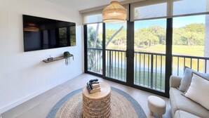A modern living room with a large TV, a round wooden table, a sofa, and a view of a tropical garden through large windows. - BT 3824 Pond View Condo - Welcome to Paradise (Fort Pierce)