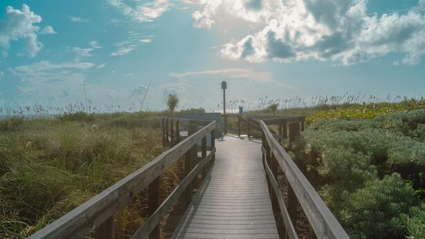 A wooden boardwalk leading to a structure with a sign, surrounded by tall grasses and shrubs. - BT 3824 Pond View Condo - Welcome to Paradise (Fort Pierce)