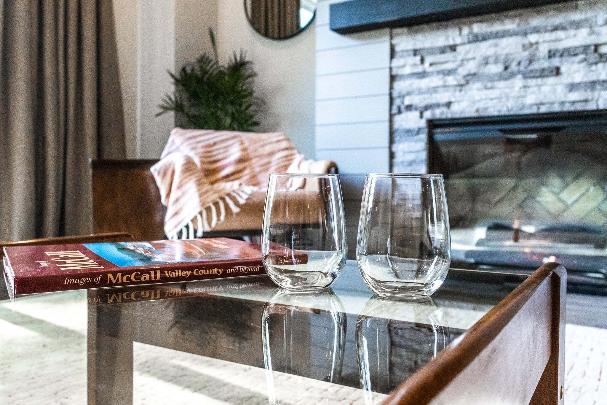 A glass table with two empty glasses, a book titled 'Images of McCall Valley County and Beyond,' and a fireplace in the background.
