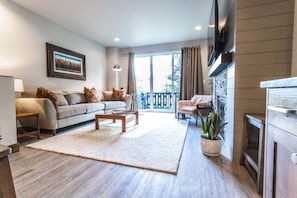 A living room with a sofa, coffee table, and a TV. There is a balcony with a railing and a plant. - Luxury in Downtown McCall, Walking Distance to Everything Downtown (McCall)