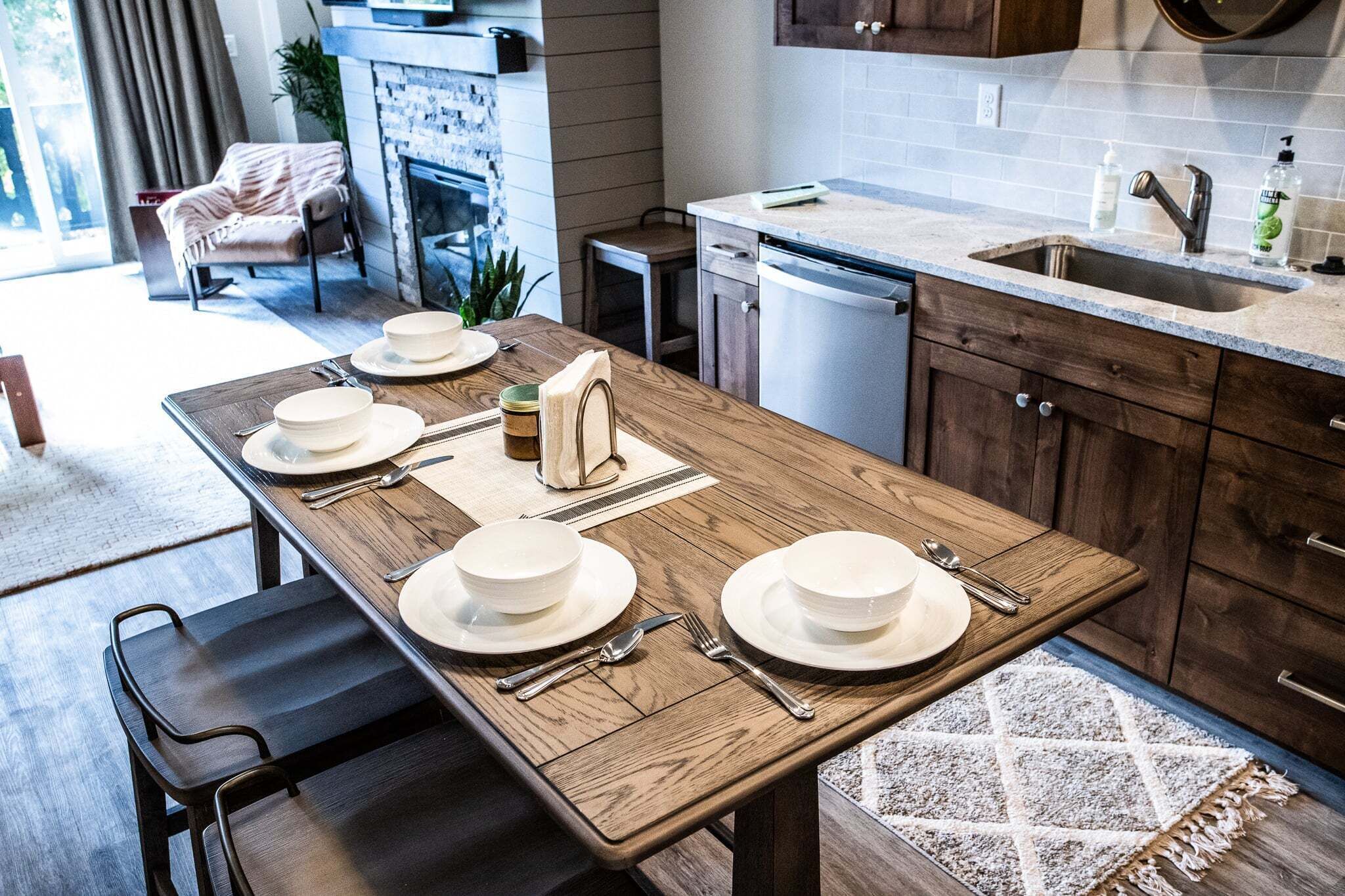 A dining area with a wooden table set for four, a kitchen with a dishwasher, and a view of the outdoors.