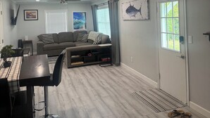 A living room with a gray sofa, a dining table with chairs, a ceiling fan, and a door with a window. - Charming 2-bedroom house with dock, AC, & Wifi. Enjoy kayaking
2 min from beach (Vero Beach)