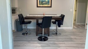 A dining area with a wooden table, four black chairs, a framed painting above the table, and a trash can to the left. - Charming 2-bedroom house with dock, AC, & Wifi. Enjoy kayaking
2 min from beach (Vero Beach)