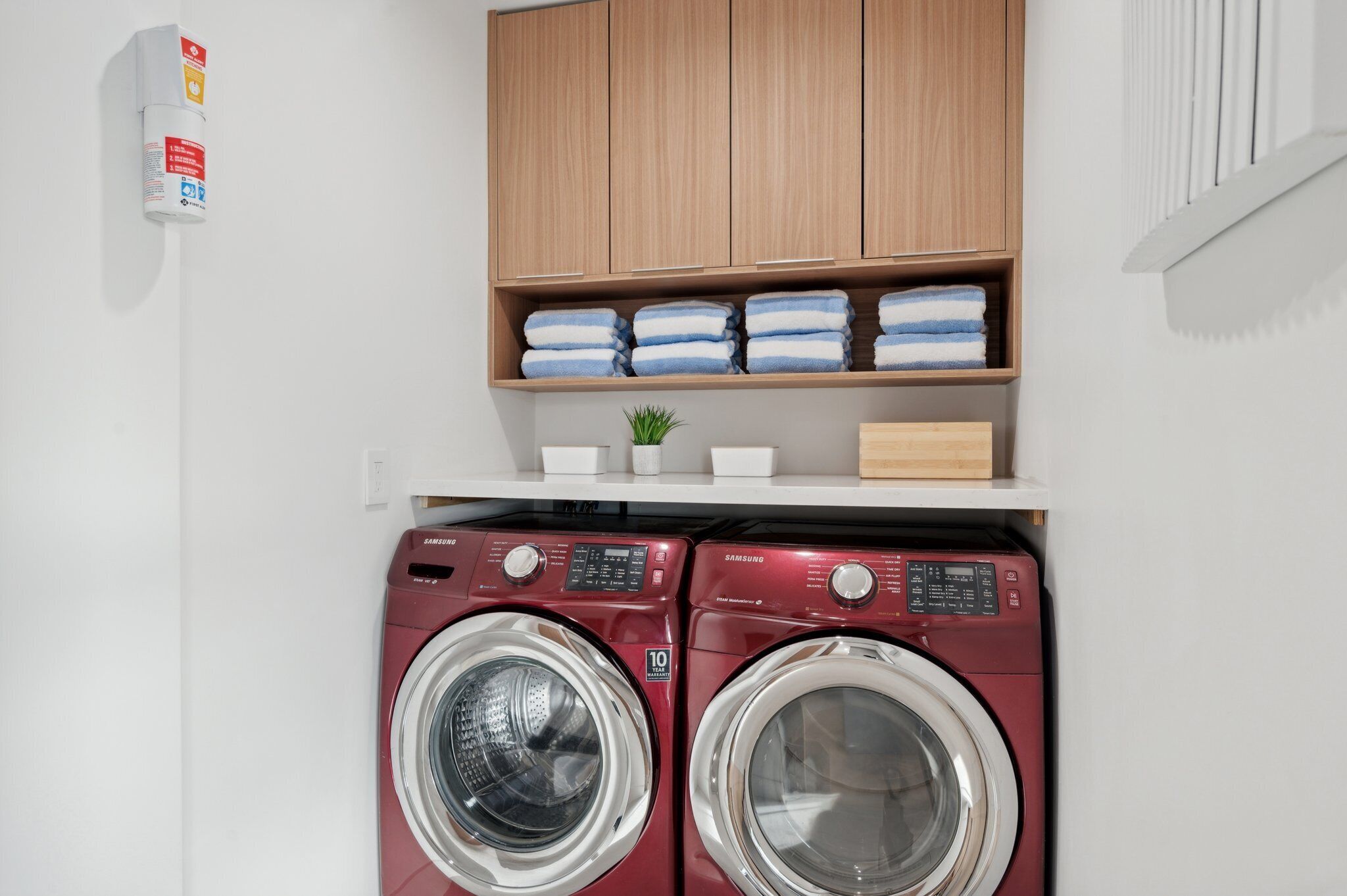 A laundry area with two red Samsung washing machines, a shelf with folded towels, and a small potted plant.