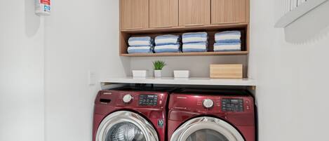 A laundry area with two red Samsung washing machines, a shelf with folded towels, and a small potted plant.