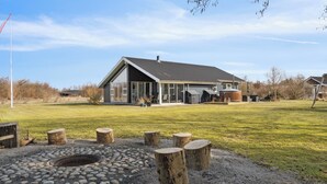 Un bâtiment moderne doté d’une grande façade en verre, d’un espace extérieur couvert et d’un foyer entouré de souches d’arbres.
