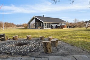 A modern building with a large glass facade, a covered outdoor area, and a fire pit surrounded by wooden stumps. - Holiday dream on the west coast (Brovst)
