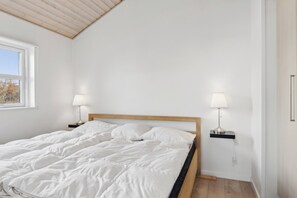 Ein Schlafzimmer mit hölzernem Bettgestell, weißer Bettwäsche, zwei Nachttischlampen und einem Fenster mit Blick auf Bäume.