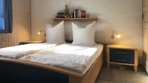 A double bed with white bedding, two bedside tables with lamps, a shelf with books and a plant, and a wooden headboard.