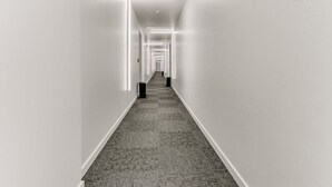 A long, narrow hallway with white walls and a carpeted floor, leading to a door at the end. - New LUX 2Bed Apt @ UCO (Edmond)